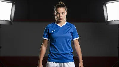 A photorealistic image of a 23-year-old female football player with a strong build and a determined expression, wearing a...