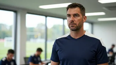 A 35-year-old male assistant coach in a navy blue and white football training kit, standing in a modern football club...