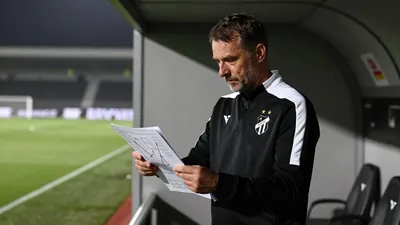 A 35-year-old Häcken coach, wearing a plain black and white polyester training top, stands in the low-light haze of the...