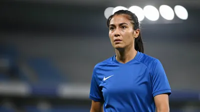 A mid-30s female right-back in a solid blue kit, waist-up, focused expression, background blurred stadium lights, sharp eyes