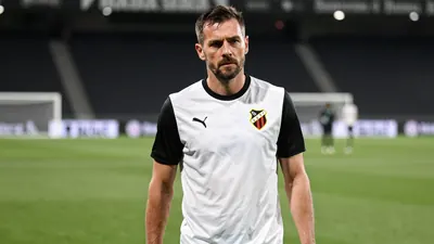 A weary, mid-30s Häcken midfielder with a strong build, dressed in a plain black and white kit, stands on the Bravida...