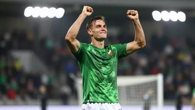 A Häcken player in a green jersey and white shorts, mid-celebration after scoring a goal, arms raised, sharp focus on the...