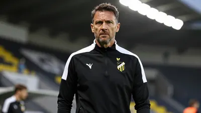 A determined, mid-30s Häcken coach with a strong build, wearing a plain black and white training top, stands pitchside at...